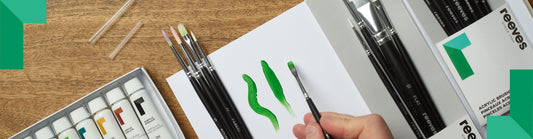 Hand painting green brushstrokes on white paper with a flat brush. Acrylic paint tubes and brushes are neatly arranged next to the painter on the table.